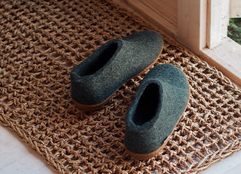 A pair of glerups shoes in the color Forest green placed on a woven doormat near a wooden doorway. The shoes have a natural rubber sole.