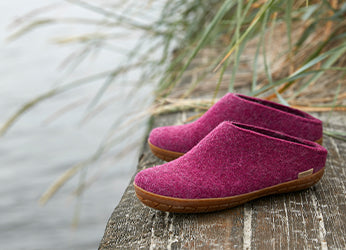 A pair of glerups slip‑ons in the color Cranberry red placed on a weathered wooden surface outdoors, with tall grass and water in the background.