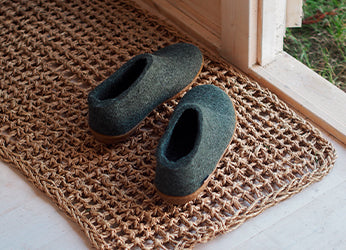 A pair of glerups shoes in the color Forest green placed on a woven doormat near a wooden doorway. 