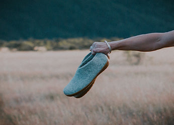 A hand holding a pair of glerups shoes in the color Grey, extended over a field with tall grass. A dark mountain ridge is visible in the background. 