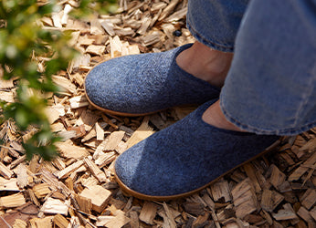 A close‑up of glerups slip‑ons in the color Denim blue worn by a person standing on wood chips.