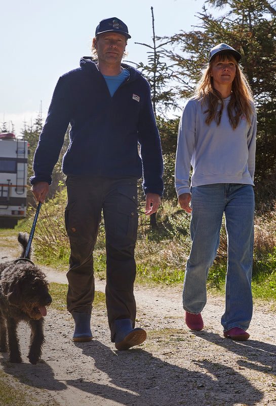 Two individuals walking on a forest path, each wearing glerups shoes. One is holding a dog’s leash, and trees and sunlight are visible in the background.