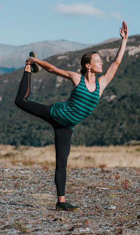 A person standing on rocky ground performing a yoga balance with one leg lifted and held behind the body. The weather is clear with blue sky and mountains in the background. The person is wearing athletic clothing and glerups shoes in the color Forest green. 