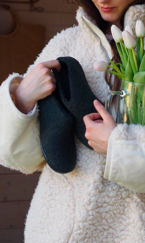 A person wearing a light, textured coat is holding a pair of Charcoal grey glerups shoes in one hand and a glass vase with light-colored tulips in the other.