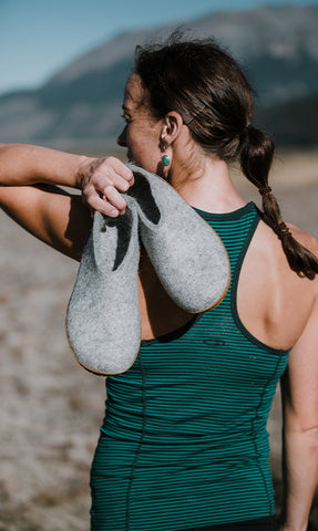 A person standing outdoors holding a pair og glerups shoes in the color grey over the shoulder. The person is wearing athletic clothing, and the background shows a natural landscape with mountains and a clear sky. 