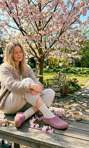 Woman sitting on a wooden garden table under a blooming cherry tree, wearing blossom pink wool slippers, surrounded by fallen blossoms and a cup of tea.