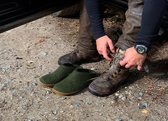 A person in a pair of Forest green glerups slip-ons sits beside worn hiking boots.