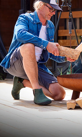 A person sitting outdoors with a peace of wood in front of a fireplace wearing glerups boots in the color Forest green with natural rubber sole in black. 