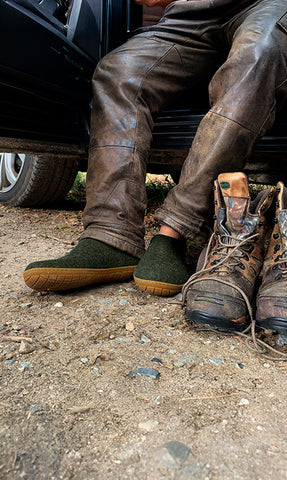 A person in a pair of Forest green glerups slip-ons sits beside worn hiking boots.