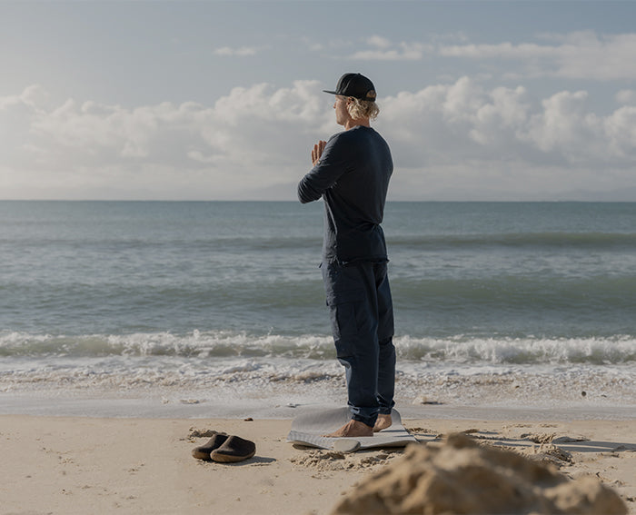 A person during yoga outdoors on a beach. A pair of glerups slip-ons are placed beside the person in the color Chestnut brown with natural rubber soles. 