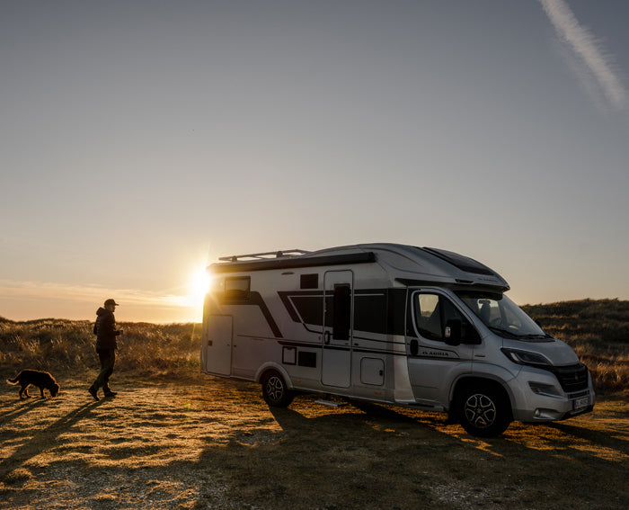 A camper van is parked in a coastal landscape at sunset, while a person walks a dog across the grassy dunes nearby.