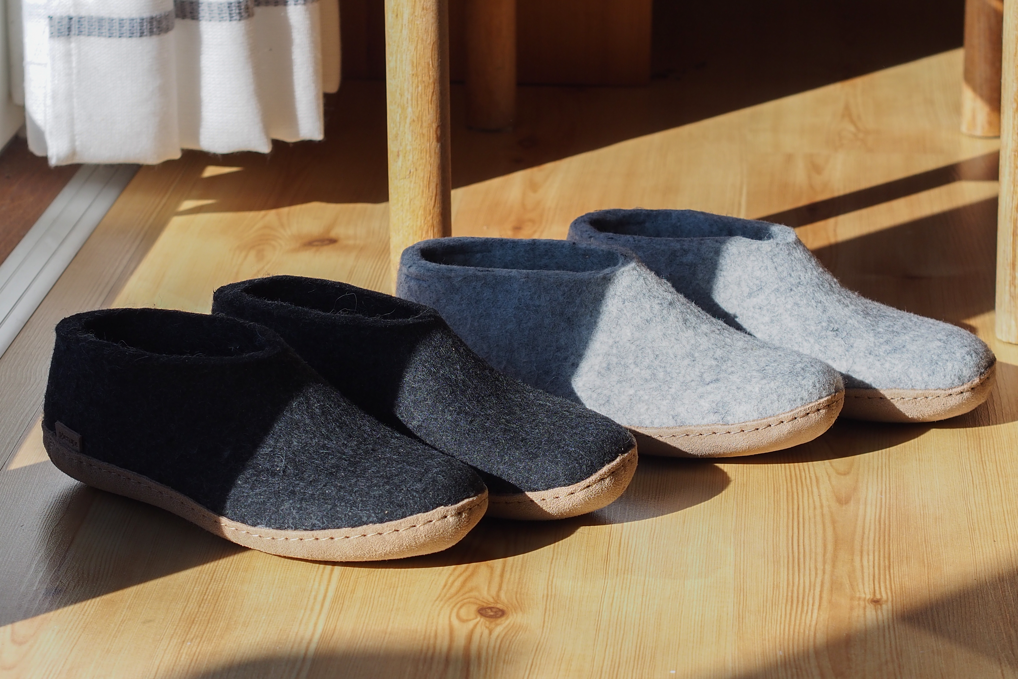 An indoor, high-angle shot of two pairs of glerups shoes sitting in a patch of bright morning sunlight on a light-colored hardwood floor near a wooden chair and a white striped curtain; one pair is Charcoal, black and the other is Grey, and both have leather soles.